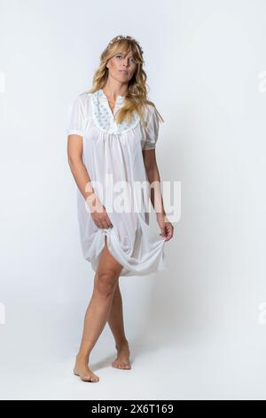 Un portrait complet du corps d'une jeune femme aux cheveux blonds ondulés, portant une robe blanche brodée et une couronne de fleurs roses. Look moderne et élégant, fashi Banque D'Images