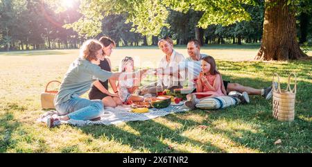 Grande famille assis sur la couverture de pique-nique dans le parc de la ville pendant le week-end journée ensoleillée. Ils souriaient, riaient et mangeaient de la pastèque, du maïs bouilli, de la tarte au col Banque D'Images