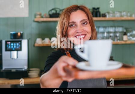 Femme barista souriante et joyeuse donnant un espresso aromatique de tasse blanche au comptoir du café. Gens heureux, consommation de café, industrie des boissons Banque D'Images