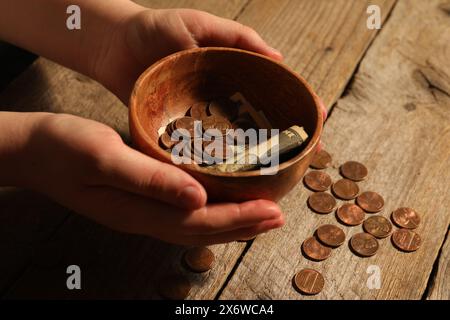 Donnez et donnez concept. Femme tenant un bol avec des pièces de monnaie et des billets de dollar à la table en bois, gros plan Banque D'Images