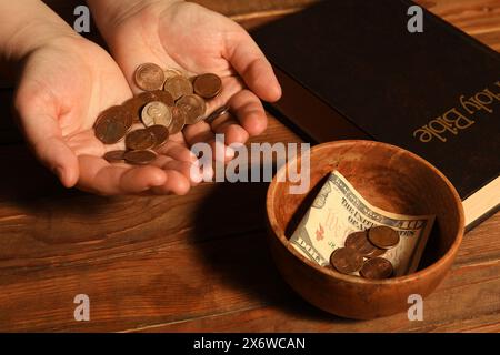 Donnez et donnez concept. Femme tenant des pièces de monnaie, gros plan. Bible et bol d'argent sur la table en bois Banque D'Images