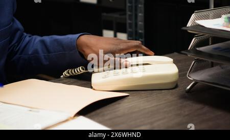 Plan détaillé d'un officier afro-américain répondant à un appel sur une ligne fixe et prenant des notes cruciales sur le cas. Expert détective masculin utilise le téléphone pour recueillir des preuves et analyser les indices dans un bureau bien équipé. Banque D'Images