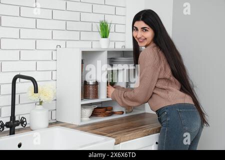 Jolie jeune femme mettant la vaisselle sur les étagères dans la cuisine légère Banque D'Images