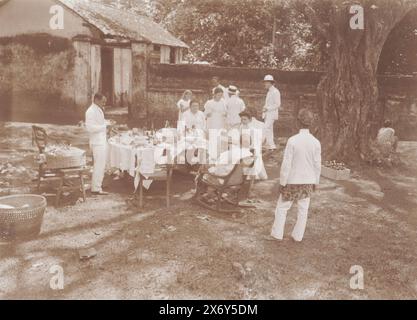Les Européens vêtus de blanc se détendent à la table extérieure tandis que le serviteur indonésien regarde de loin., Batuwan près de Soemenep 1910 (titre sur l'objet), photographies (titre de la série), les hommes portent des uniformes tropicaux blancs, un homme avec un casque à moelle, deux autres hommes portent un chapeau de paille, les femmes portent des robes blanches ou des jupes en batik avec un gilet blanc. Photo tirée de l'album 'photographies'., photographie, Frits Freerks Fontein Fz., (attribué à), Frits Freerks Fontein Fz., (attribué à), Madura, (éventuellement), pays-Bas, (éventuellement), c. 1910, carton, hauteur, 79 mm × largeur, 109 mm, hauteur, 242 mm × largeur Banque D'Images