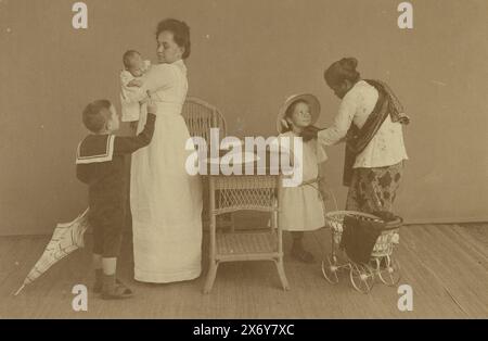 Trois enfants inconnus avec leur mère et leur gardienne indonésienne (appelée « baboue » à l'époque). À gauche d'une table et d'une chaise se trouve une femme debout dans une robe blanche avec un bébé dans son bras. À côté d'elle un garçon en costume de marin avec un parapluie donnant au bébé une sucette. Sur la droite, une fille avec une poussette de poupée qui est mise sur un chapeau par une gardienne indonésienne (appelée 'baboue' à l'époque) dans un sarong et kabaja., photographie, anonyme, Indes orientales néerlandaises, c. 1905 - c. 1925, papier baryta, impression gélatineuse argentée, hauteur, 115 mm × largeur, 170 mm Banque D'Images