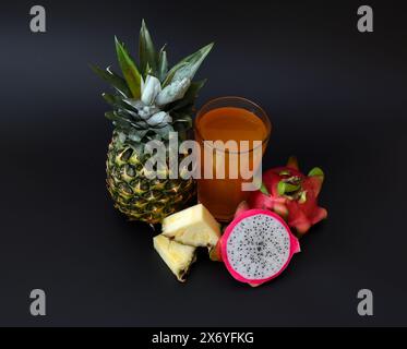 Un grand verre de jus jaune fruité sur un fond noir avec des morceaux d'ananas mûr et un demi-pitaya mûr. Gros plan. Banque D'Images