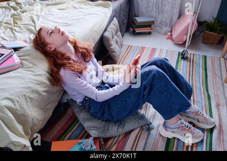 Vue en angle élevé d'une adolescente mélancolique avec des cheveux lus tenant un smartphone assis sur le sol dans sa chambre regardant vers le haut au plafond Banque D'Images