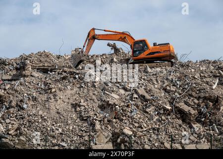 Excavatrice orange opérant sur un gros tas de gravats et de débris sur un site de démolition sous un ciel nuageux. Banque D'Images