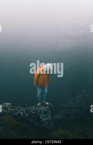 Bulgarie - 05 22 2024 : homme avec une veste orange et un Jean bleu debout sur le bord d'un grand rocher regardant vers le bas de la falaise avec un b sombre et brumeux et pluvieux Banque D'Images