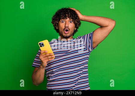 Portrait de gars aux cheveux bouclés en t-shirt rayé à l'aide de la tête tactile de smartphone confondu avec des choses Internet isolées sur fond de couleur verte Banque D'Images