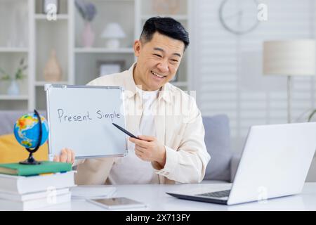 Professeur de langue en ligne souriant expliquant le temps simple présent au cours d'une leçon virtuelle. Homme tenant un tableau blanc et pointant avec un stylo tout en enseignant via un ordinateur portable. Banque D'Images