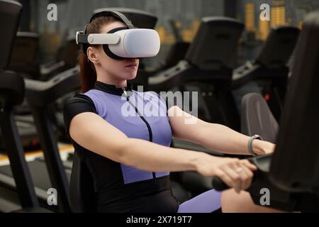 Portrait d'une jeune femme portant un casque VR dans le gymnase et utilisant un vélo d'exercice dans la simulation d'entraînement immersive Banque D'Images