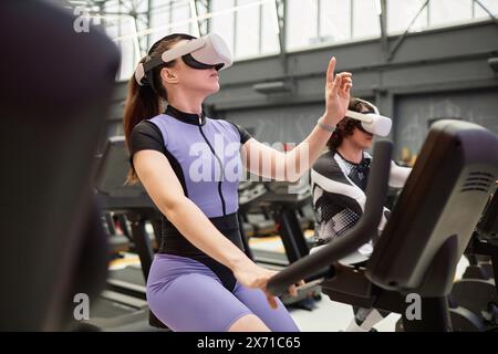 Portrait de vue latérale de la jeune femme utilisant la technologie VR immersive tout en s'entraînant sur le vélo d'exercice dans l'espace de copie futuriste de gymnastique Banque D'Images