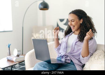 Heureuse jeune femme aux cheveux bouclés brésilienne ou hispanique excitée, freelance ou web designer, assise sur un canapé confortable avec ordinateur portable, se réjouissant du succès, faisant des gestes avec les poings, souriant Banque D'Images
