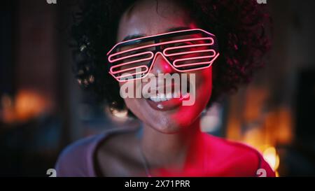 Gros plan Portrait de jeune femme latine noire multiethnique avec des cheveux afro dans des lunettes futuristes néon luisantes, s'amuser à la maison, sourire et poser pour la caméra dans l'appartement loft dans la soirée. Banque D'Images