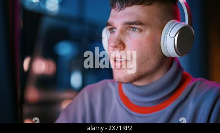 Gros plan Portrait de bel homme dans les écouteurs à l'aide de l'ordinateur dans élégant appartement loft la nuit. Homme créatif travaillant à domicile, naviguant sur Internet et les médias sociaux. Vue sur la ville urbaine depuis la grande fenêtre. Banque D'Images