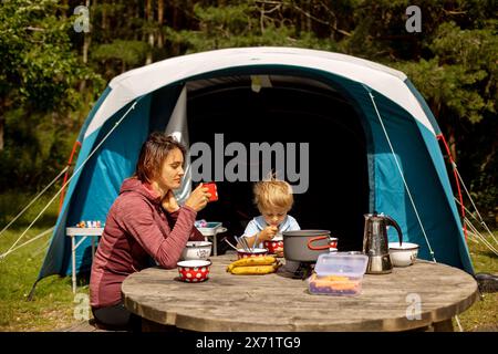 Famille, mère et enfant, prenant le petit déjeuner devant une tente dressée dans la forêt, tandis que camping sauvage en Norvège, en été Banque D'Images