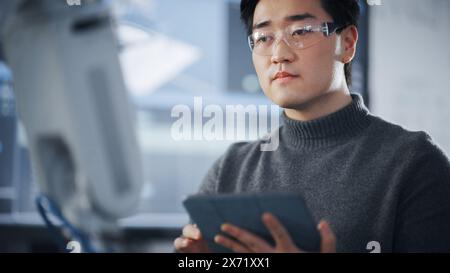 Étudiant utilisant la tablette pour contrôler la main robotique. Bras bionique se déplaçant dans différentes directions pendant la classe universitaire. Homme asiatique développant un logiciel pour l'installation de robotique. Concept informatique. Banque D'Images