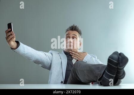 Homme d'âge moyen portant un blazer gris clair, chemise gris foncé et cravate rayée, prend un selfie avec un bras tendu, tandis que ses pieds reposent sur un blanc Banque D'Images