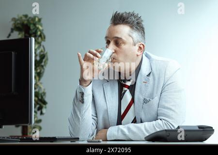 Homme avec les cheveux gris piquants, blazer gris clair, chemise gris foncé et cravate rayée, tient un verre d'alcool avec un regard souillé, croyant qu'il est normal de boire Banque D'Images