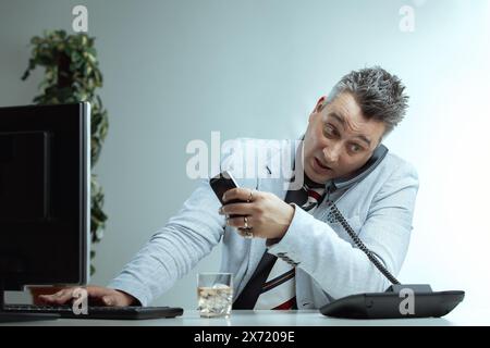 Homme avec des cheveux gris piquants, veste de costume gris clair, chemise gris foncé et cravate rayée, tient en toute confiance un verre d'alcool avec une expression maculée, Seate Banque D'Images