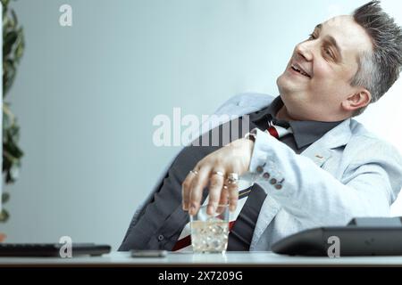 Homme d'âge moyen en blazer gris clair, chemise gris foncé, et cravate rayée, avec des cheveux gris piquants, tient un verre d'alcool avec un regard souillé, pensant qu'il n Banque D'Images