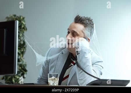 Homme aux cheveux gris piquants, veste de costume gris clair, chemise gris foncé et cravate rayée, couvert de toiles d'araignée, parle au téléphone avec une expression fatiguée, dr. Banque D'Images