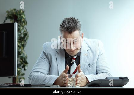 Homme caucasien en blazer gris clair, chemise gris foncé, et cravate rayée, avec des cheveux gris piquants, tient en toute confiance un verre d'alcool, montrant un expre souillé Banque D'Images