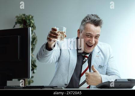 Smug homme dans une veste de costume gris clair, chemise gris foncé, et cravate rayée, avec des cheveux gris piquants, tient en toute confiance un verre d'alcool, assis à un bureau, Banque D'Images