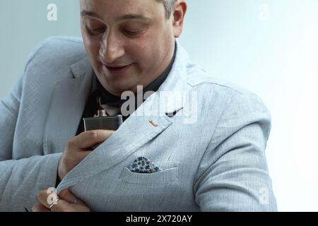 Homme caucasien en blazer gris clair, chemise gris foncé, et cravate rayée, avec des cheveux gris piquants, sirote nerveusement d'un flacon tout en regardant sur le côté, feari Banque D'Images