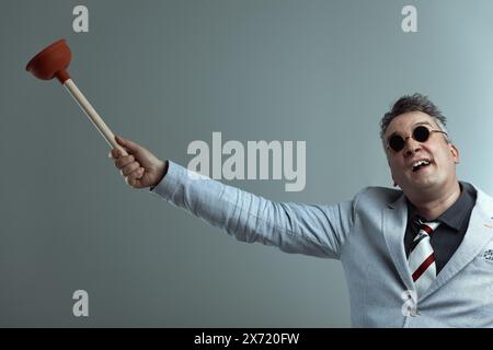 Homme en veste de costume gris clair, chemise gris foncé, et cravate rayée, portant des lunettes de soleil rondes, tient un plongeur haut avec une expression joyeuse contre un Banque D'Images