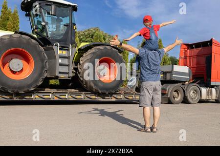 un homme et un garçon assis sur ses épaules se tiennent devant un grand tracteur chargé sur une remorque de camion. la passion de son pour les voitures. Fête des pères, sois ensemble. enfant Banque D'Images