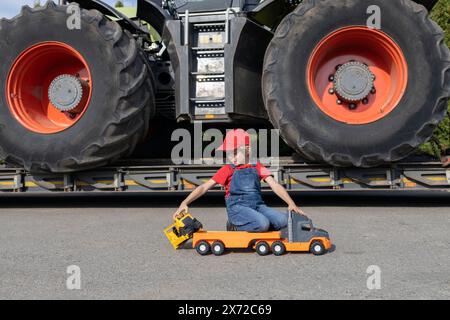 garçon joue avec des voitures jouets. Derrière lui se trouve un vrai tracteur avec d'énormes roues. L'enfant veut devenir chauffeur ou mécanicien, comme papa. vehic commercial Banque D'Images