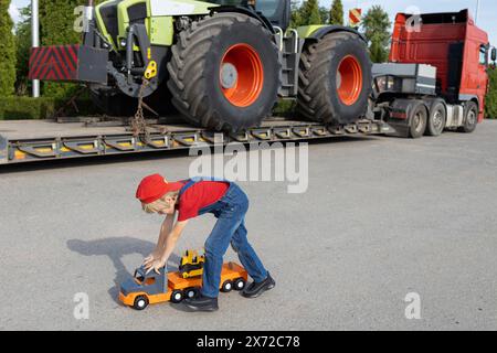 garçon joue avec des voitures jouets dans le parking à côté d'un vrai gros camion avec un tracteur chargé. L’enfant rêve de devenir chauffeur ou mécanicien, comme papa Banque D'Images