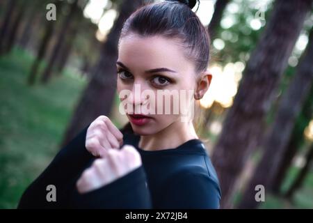 Jeune femme faisant la formation d'arts martiaux dans le haut noir sportif tenant les mains dans la pose de combat sur fond de forêt. Banque D'Images