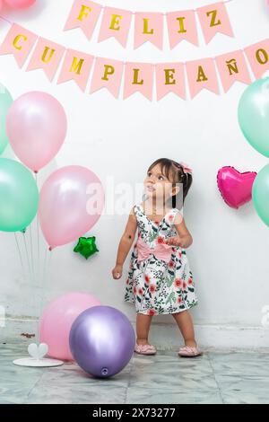 petite brune latina fille levant les yeux tout en agitant ses mains et dansant le jour de son anniversaire entourée de ballons Banque D'Images