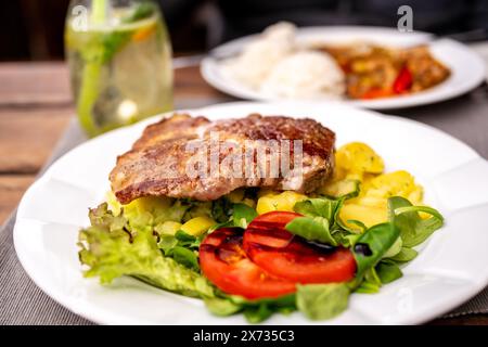 Steak de porc sur salade de pommes de terre avec légumes sur la première assiette blanche, boire dans le verre et la deuxième assiette avec riz, viande de poulet et poivron rouge sur fond o Banque D'Images