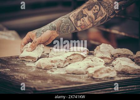 Un boulanger avec des tatouages met des petits pains de pâte crus sur la plaque de cuisson dans la boulangerie Banque D'Images