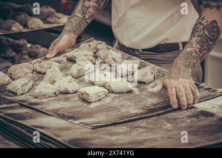 Un boulanger avec des tatouages met des petits pains de pâte crus sur la plaque de cuisson dans la boulangerie, style vintage avec du grain Banque D'Images