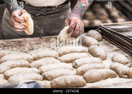 Un boulanger avec des tatouages Un boulanger avec des tatouages met des petits pains de pâte crus sur la plaque de cuisson dans la boulangerie. Banque D'Images