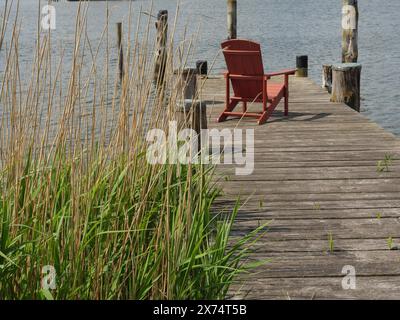 Une jetée en bois tranquille sur le bord du lac avec un seul siège rouge et des roseaux verts au premier plan, jetée en bois avec une chaise et un filet de pêche par le Banque D'Images