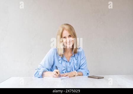 Belle femme souriante mi-adulte écrit dans son cahier tout en regardant la caméra Banque D'Images