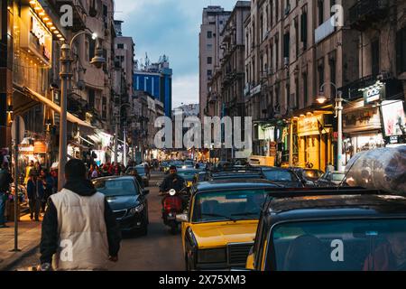 Scène de rue animée à Alexandrie, en Égypte, avec un trafic dense avec des taxis jaunes, des piétons et des vitrines éclairées au crépuscule Banque D'Images