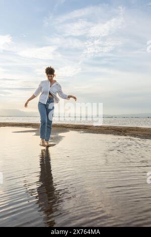 femme sur la plage dansant au coucher du soleil en contre-jour avec le soleil derrière dans l'ombre, femme en vacances marchant sur le sable avec des reflets d'eau dorée f Banque D'Images