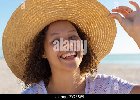 À la plage, jeune femme biraciale tenant un chapeau à large bord, riant Banque D'Images