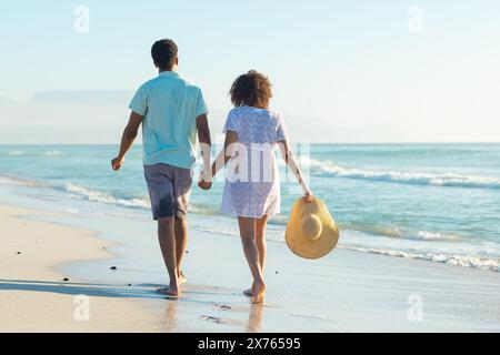 À la plage, couple biracial se tenant la main, marchant le long du rivage Banque D'Images