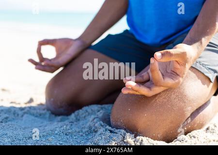 A la plage, jeune homme biracial assis, méditant en chemise bleue Banque D'Images