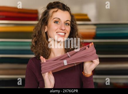 Créatrice de mode enthousiaste affichant fièrement un accessoire textile fraîchement fabriqué dans son studio bien organisé, entouré de rouleaux de tissus colorés Banque D'Images