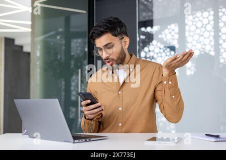 Employé de bureau qui semble frustré lorsqu'il lit de mauvaises nouvelles sur son téléphone tout en travaillant sur son ordinateur portable. Banque D'Images
