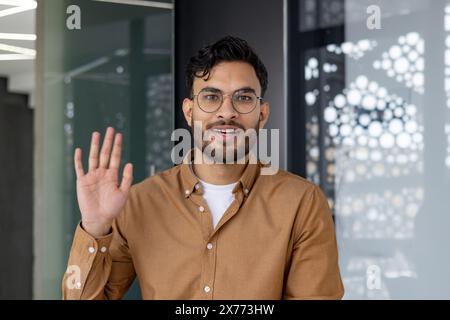 Jeune homme agitant la main et regardant la caméra pendant un appel vidéo dans un bureau moderne. Concept de communication professionnelle, de réunion virtuelle et de conversation en ligne. Banque D'Images